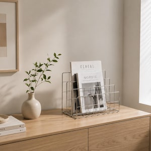 Può includere: Un portariviste in filo metallico color argento dal design a gradini, contenente due riviste intitolate CEREAL e NORDIC. Il supporto presenta una base rettangolare e divisori verticali di altezze variabili realizzati con sottili aste.