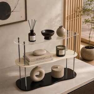 Könnte beinhalten: Ein dreistufiges Regal mit verchromten Metallstützen und wellenförmigen Ablagen in transparentem Acryl, Creme und Schwarz. Die Ablagen enthalten einen Diffusor, eine schwarze Schale, eine beige Vase, zwei Bücher mit den Titeln Eat Drink Nap und Kinfolk, eine Kerze, eine ringförmige Vase und einen gerippten Behälter mit Deckel.