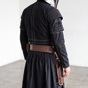 Op de afbeelding: Een metallic zilveren helm met een donker vizier gecombineerd met een zwart jasje met witte contraststiksels op de schouders en rug. Een bruine lederen utility-riem met holster wordt gedragen over een lange zwarte rok met zijsplit.