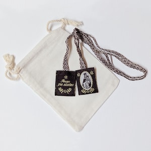 May include: A brown fabric scapular featuring a woven cord and two rectangular panels. One panel displays an oval image of a figure holding a child, while the other panel features the gold embroidered text "Ruega por nosotros" with floral accents, accompanied by a small off-white drawstring pouch.
