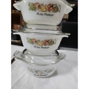 May include: A stack of three white milk glass casserole dishes with clear glass lids. Each dish features a vegetable illustration in orange, green, and yellow tones with the text Petite Potager printed in black lettering on the front side.