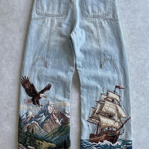 Könnte beinhalten: Hellblaue Jeans aus Denim mit einem schwarzen Markenetikett am hinteren Bund. Die Hosenbeine zeigen detaillierte Stickereien: einen Weißkopfseeadler über einem Gebirge mit Tannen auf der linken Seite und ein Segelschiff auf Wellen rechts.