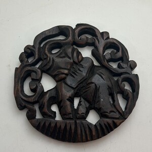 May include: A dark brown, circular wooden carving of an elephant. The elephant is surrounded by ornate, swirling patterns. The carving is detailed and appears to be hand-carved, showcasing the elephant's features and the decorative border.