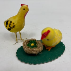 May include: Two yellow felt chicks with red combs, one standing and one sitting next to a small woven nest with a yellow egg inside. The nest is on a green felt oval.