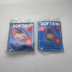 May include: Two packages of Softies craft kits, each featuring a different character. The first package shows a red and white character with a black hat and a red cape. The second package shows a blue and white character with a purple hat and a blue cape. Both packages have the text "Softies" printed on them.