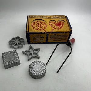 May include: A vintage double rosette and timbale iron set featuring a yellow and blue cardboard box with printed illustrations, four distinct metal molds in flower, star, square, and round shapes, and a metal handle with a red wooden grip.