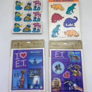 Könnte beinhalten: Eine Sammlung von Vintage-Hallmark-Aufklebern. Die Aufkleber zeigen Charaktere aus der Sesamstraße, Dinosaurier und dem Film E.T. Der Außerirdische. Die Sesamstraßen-Aufkleber haben die Wörter "Good" und "Much Better". Die E.T.-Aufkleber haben den Text "I Love E.T."