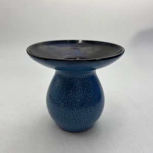 May include: A small, blue ceramic vase with a speckled glaze. The vase has a wide, flat top and a narrow base.