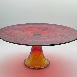 May include: A red glass cake stand with a scalloped edge and a yellow glass base. The stand is decorated with a grapevine pattern.