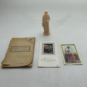 May include: A collection of religious items, including a beige figurine of a saint, a vintage book titled "Saint Joseph," and two religious cards with illustrations. The items are arranged on a light surface.