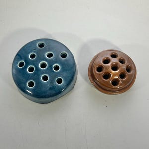 May include: Two ceramic flower frogs. One is a round, blue-green color with multiple holes for arranging stems. The other is a smaller, round, brown frog with similar holes. These are used for floral arrangements.