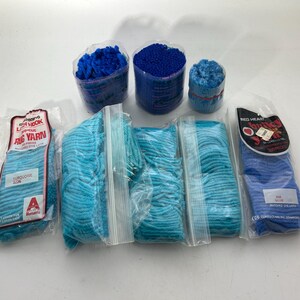 Könnte beinhalten: Sortiment an blauen Garnknäueln und -kugeln, darunter Garn der Marke Red Heart, Garn der Marke Lion Brand und ein Knäuel türkisfarbenes Garn. Das Garn ist in Plastiktüten verpackt und bereit zum Verarbeiten.