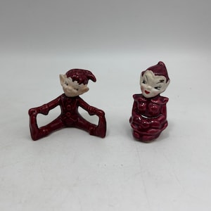 May include: Two ceramic elf figurines, one sitting and one in a seated position with legs outstretched. Both are painted in a deep burgundy color with white facial features.