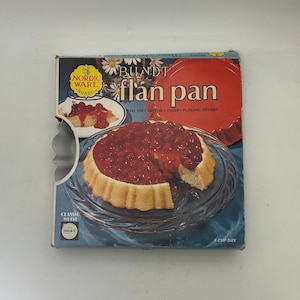 May include: A blue and red box containing a white flan pan. The box features the text "Bundt Flan Pan" and "Nordic Ware". The pan is 4 cup size and is made from classic white Teflon.