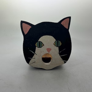 Puede incluir: Un objeto decorativo de madera con forma de gato. El gato tiene pelaje negro con marcas blancas, orejas rosas y ojos verdes. La boca está abierta, revelando un interior marrón. El artículo está pintado y parece hecho a mano.