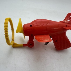Puede incluir: Un avión de juguete de plástico rojo con una hélice amarilla y un anillo amarillo unido al lateral. El avión tiene un gatillo negro y un mango rojo.