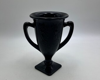 Peut inclure: Un vase en verre noir avec deux anses et un motif gravé. Le vase a une base carrée et un corps rond.