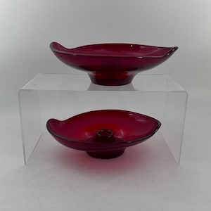 Puede incluir: Dos candelabros de vidrio rojo, uno apilado sobre el otro. El candelabro superior es un plato poco profundo con un borde ligeramente acampanado. El candelabro inferior es un plato más profundo con un portavelas central.