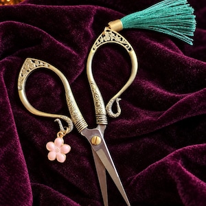 May include: Antique-style metal scissors featuring ornate, engraved handles with a brass finish. A small, light pink five-petaled flower charm hangs from one handle, and a vibrant teal green tassel is attached to the top of the other handle.