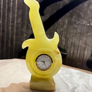 May include: A yellow, translucent stone clock with a flame-like shape mounted on a rectangular base. The clock face is circular with a gold-colored rim, black numerals from 1 to 12, and three thin black hands. The word Quartz is printed on the face.