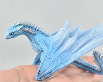 Op de afbeelding: Een klein, blauw drakenbeeldje met zilveren accenten. De draak heeft uitgespreide vleugels en een lange staart. Het beeldje is gemaakt van een getextureerd materiaal en lijkt handgeschilderd te zijn.