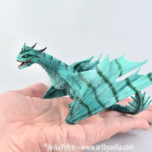 May include: A teal green dragon figurine with dark green stripes on its wings and body. The dragon has a long neck and tail, and its wings are spread out.