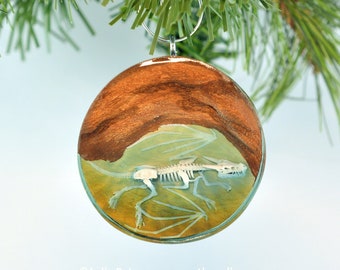 May include: A round, wooden Christmas bauble with a clear resin dome containing a detailed, white skeletal dragon with wings. The dragon is set against a green and brown background.