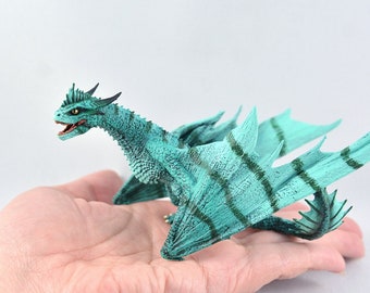 Op de afbeelding: Een tealgroene drakenfiguur met donkergroene strepen op zijn vleugels en lichaam. De draak heeft een lange nek en staart, en zijn vleugels zijn uitgespreid.