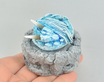 Könnte beinhalten: Eine kleine, blaue und weiße Eisdrachenfigur mit weißen Hörnern, die auf einem grauen Felssockel sitzt.
