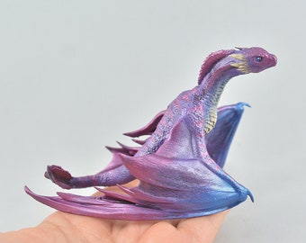 Op de afbeelding: Een klein, paars en blauw drakenbeeldje met gouden accenten. De draak heeft zijn vleugels uitgespreid en staat in een vliegende houding.