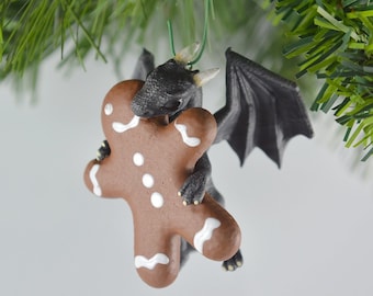 May include: A black dragon ornament with wings outstretched, holding a gingerbread man cookie. The dragon has yellow eyes and a white frosting design on the cookie.