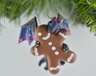 May include: A small, purple and black dragon ornament with galaxy-like wings is holding a gingerbread cookie. The ornament is hanging from a green string.