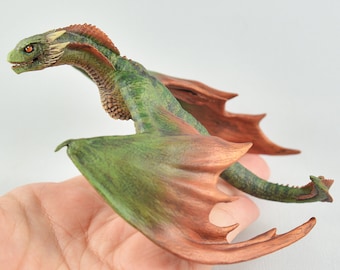 May include: A small, green and brown dragon figurine with outstretched wings. The dragon has a long neck and a pointed tail. The figurine is made of polymer clay and is painted with realistic details.