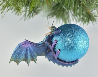 May include: A blue and purple dragon ornament clinging to a sparkly blue Christmas ball. The dragon has detailed wings and horns, and the ornament hangs from a green string, perfect for holiday decor.