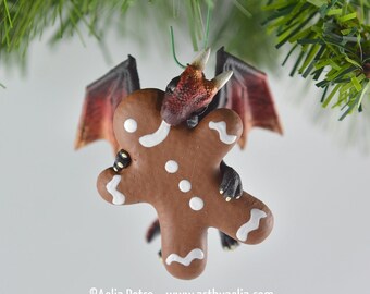 May include: A whimsical Christmas ornament featuring a dragon holding a gingerbread man. The dragon has red and brown scales, black claws, and red wings. The gingerbread man is brown with white icing details. The ornament hangs from a green wire.