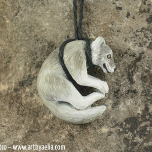 May include: A hand-carved wooden pendant of a curled up wolf, painted in shades of gray and white. The wolf has a black cord attached to it.