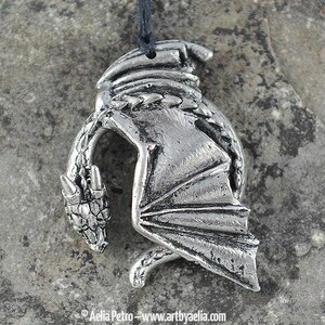 May include: Silver pendant necklace featuring a detailed dragon design. The dragon is curled up with its wings spread out. The pendant is made of pewter and has a black cord.