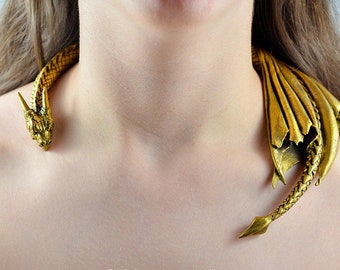 May include: A gold-colored dragon necklace with a detailed head and wings. The necklace is worn around a person's neck.