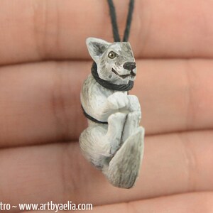 May include: A small, hand-carved wooden wolf pendant with a gray and white paint scheme. The wolf is wearing a black collar and has a friendly expression.