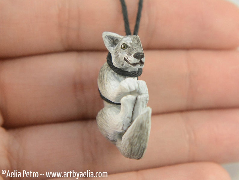 May include: A small, hand-carved wooden wolf pendant with a gray and white paint scheme. The wolf is wearing a black collar and has a friendly expression.
