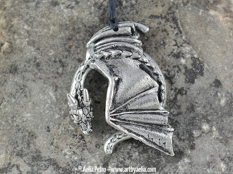 May include: Silver pendant necklace featuring a detailed dragon design. The dragon is curled up with its wings spread out. The pendant is made of pewter and has a black cord.