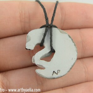 May include: White ceramic pendant necklace with a stylized animal design. The pendant is shaped like a curled-up animal with a small hole in the center. The necklace is made of black cord. The pendant has the initials "NP" inscribed on the bottom.