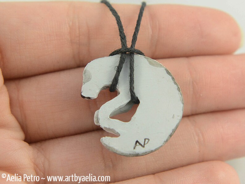 May include: White ceramic pendant necklace with a stylized animal design. The pendant is shaped like a curled-up animal with a small hole in the center. The necklace is made of black cord. The pendant has the initials "NP" inscribed on the bottom.