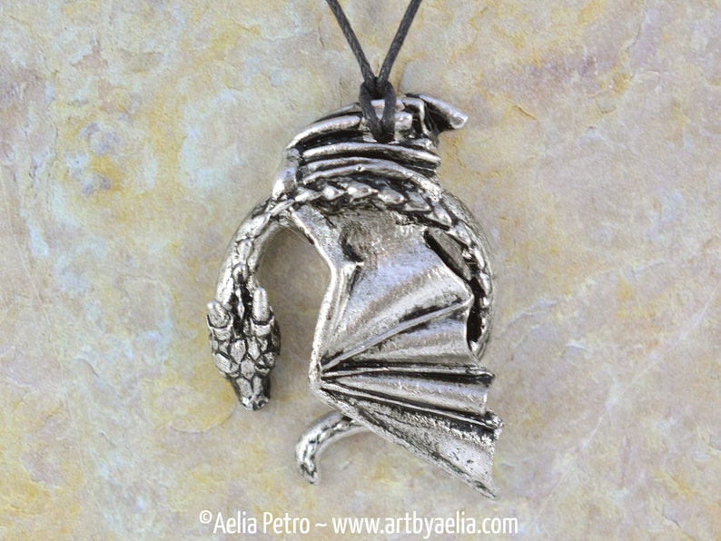 May include: Silver pendant necklace featuring a detailed dragon design with wings outstretched. The dragon is curled in a defensive posture with its head turned back towards its tail.
