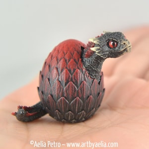 May include: A small, black and red dragon sculpture emerging from a red and black egg. The dragon has red eyes and a detailed, textured body.