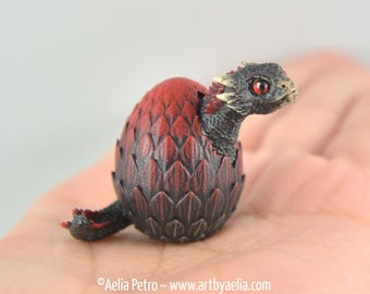 Op de afbeelding: Een klein, zwart en rood drakenbeeld dat uit een rood en zwart ei komt. De draak heeft rode ogen en een gedetailleerd, getextureerd lichaam.