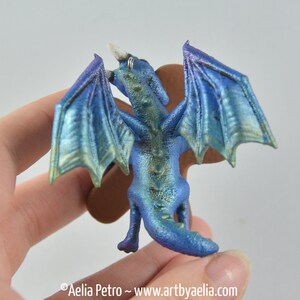 May include: A miniature blue dragon figurine with iridescent wings and a horn. The dragon is held in a hand, showcasing its detailed scales and posture. The wings have a gradient of blue and green hues.