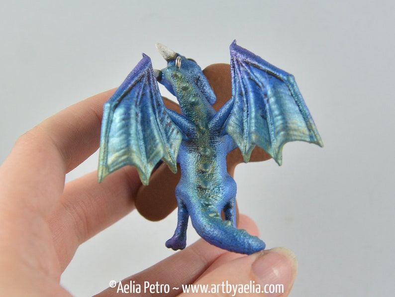 May include: A miniature blue dragon figurine with iridescent wings and a horn. The dragon is held in a hand, showcasing its detailed scales and posture. The wings have a gradient of blue and green hues.
