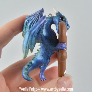 May include: A blue and purple dragon figurine with iridescent wings and a brown pole. The dragon has a small loop for hanging. The dragon is holding a brown pole with white dots. The dragon is a fantasy-themed item.