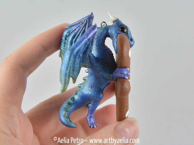 May include: A blue and purple dragon figurine with iridescent wings and a brown pole. The dragon has a small loop for hanging. The dragon is holding a brown pole with white dots. The dragon is a fantasy-themed item.